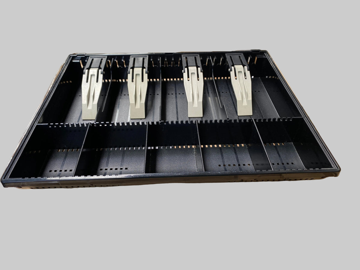 A black plastic multi-compartment organizer tray with six white plastic inserts, shown resting on top of cardboard shipping boxes in a warehouse setting.