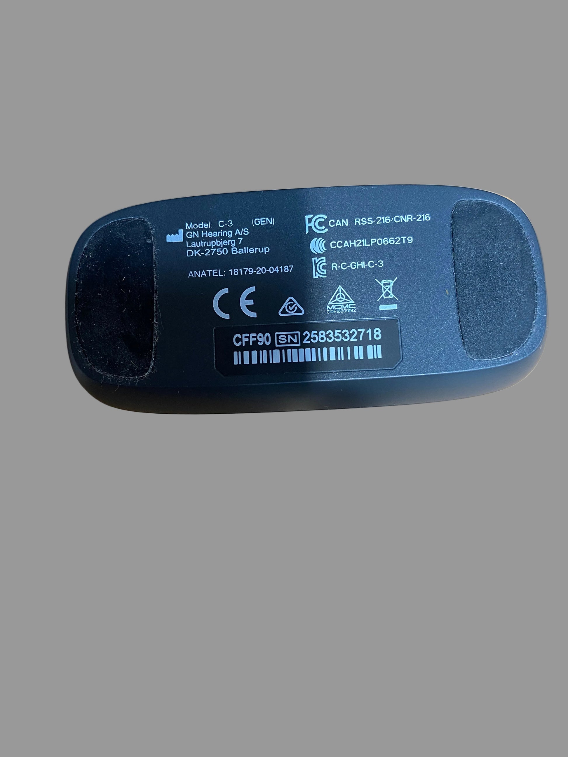 A close-up shot of the bottom of a black plastic electronic device, showing printed specifications, barcodes, and regulatory marks, resting on a brown cardboard surface.